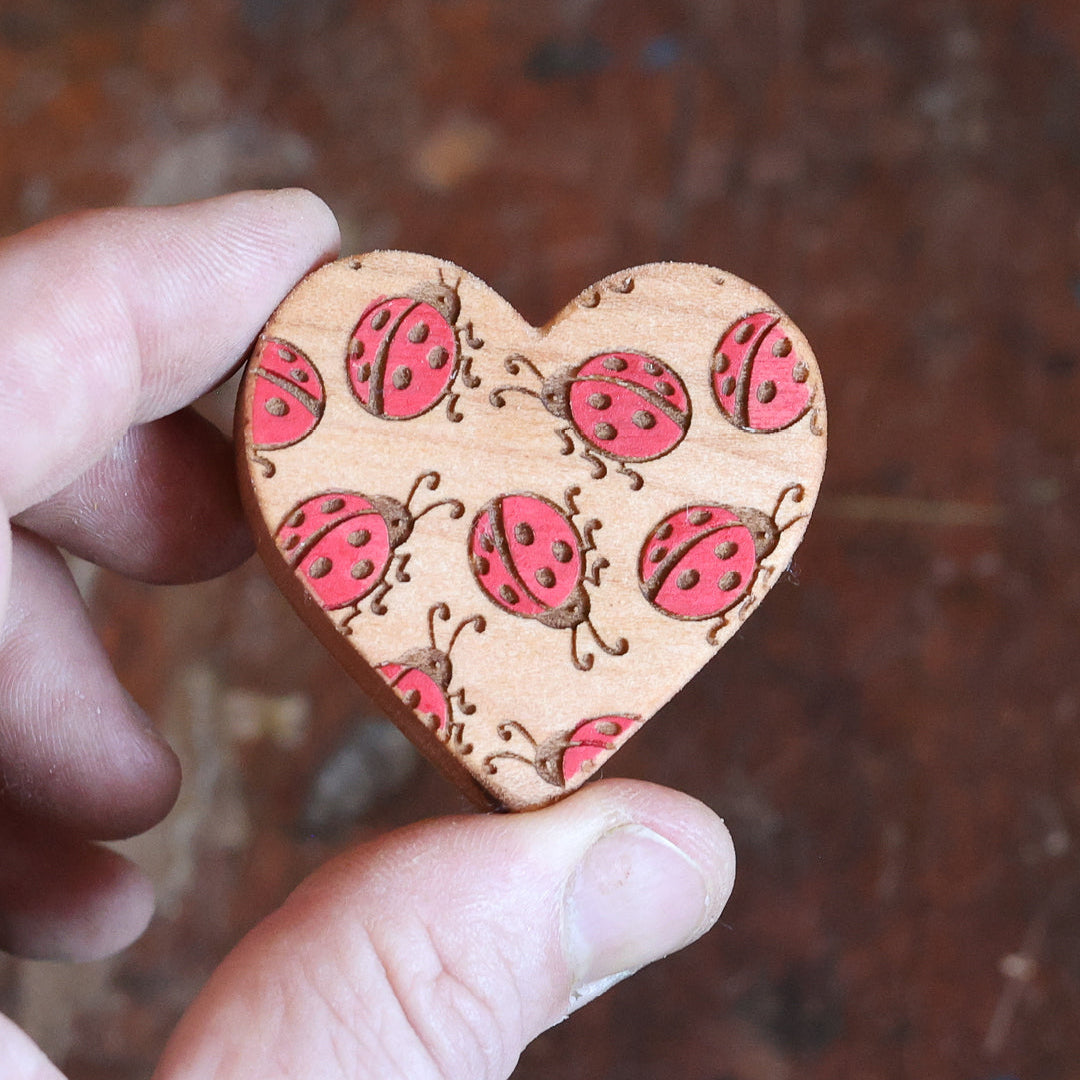 A heart-shaped wooden charm featuring a repeating pattern of red ladybugs with black spots, held between two fingers against a dark, rustic brown background.