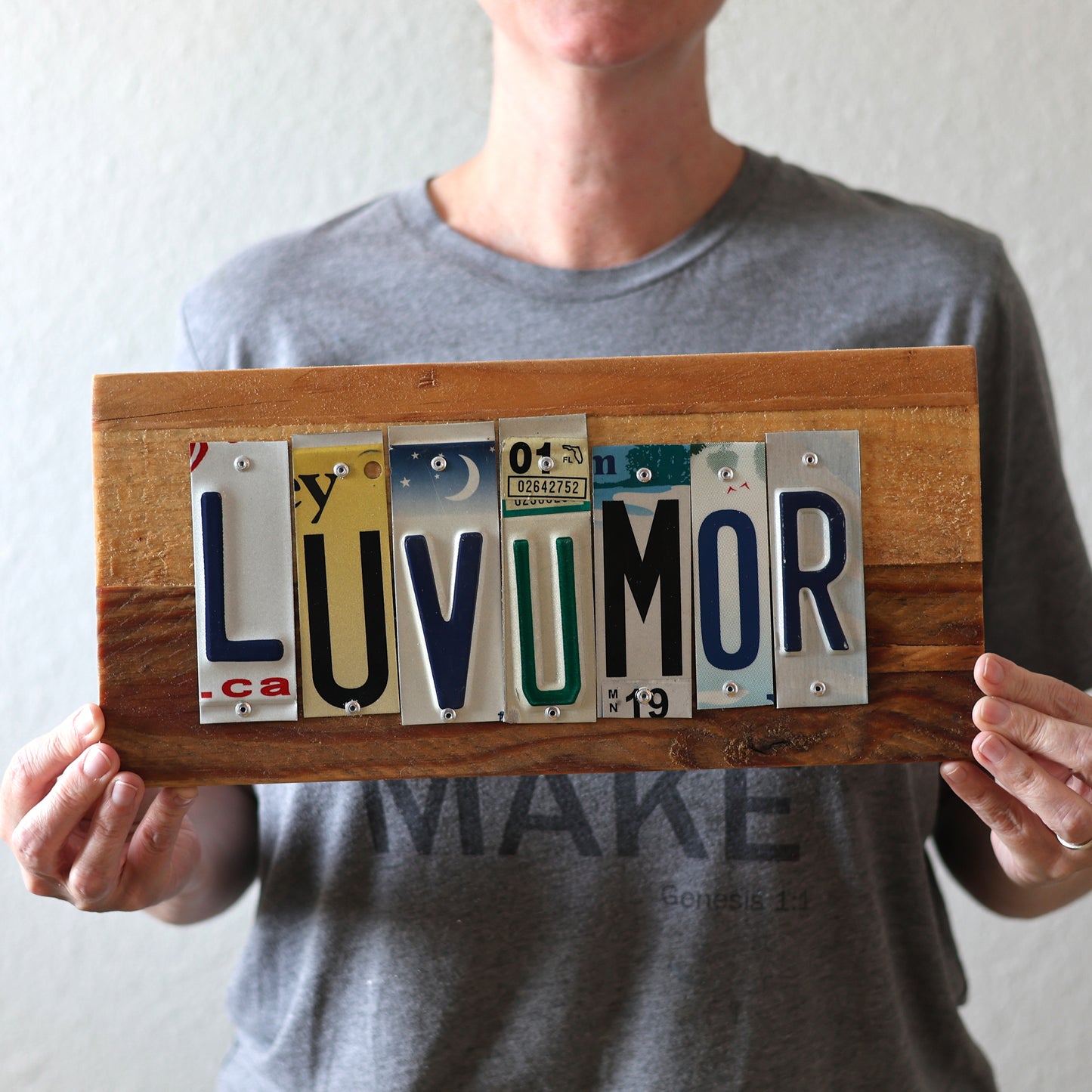 Person holding a wooden sign with license plate letters spelling 'LUVUMOR' against a plain background