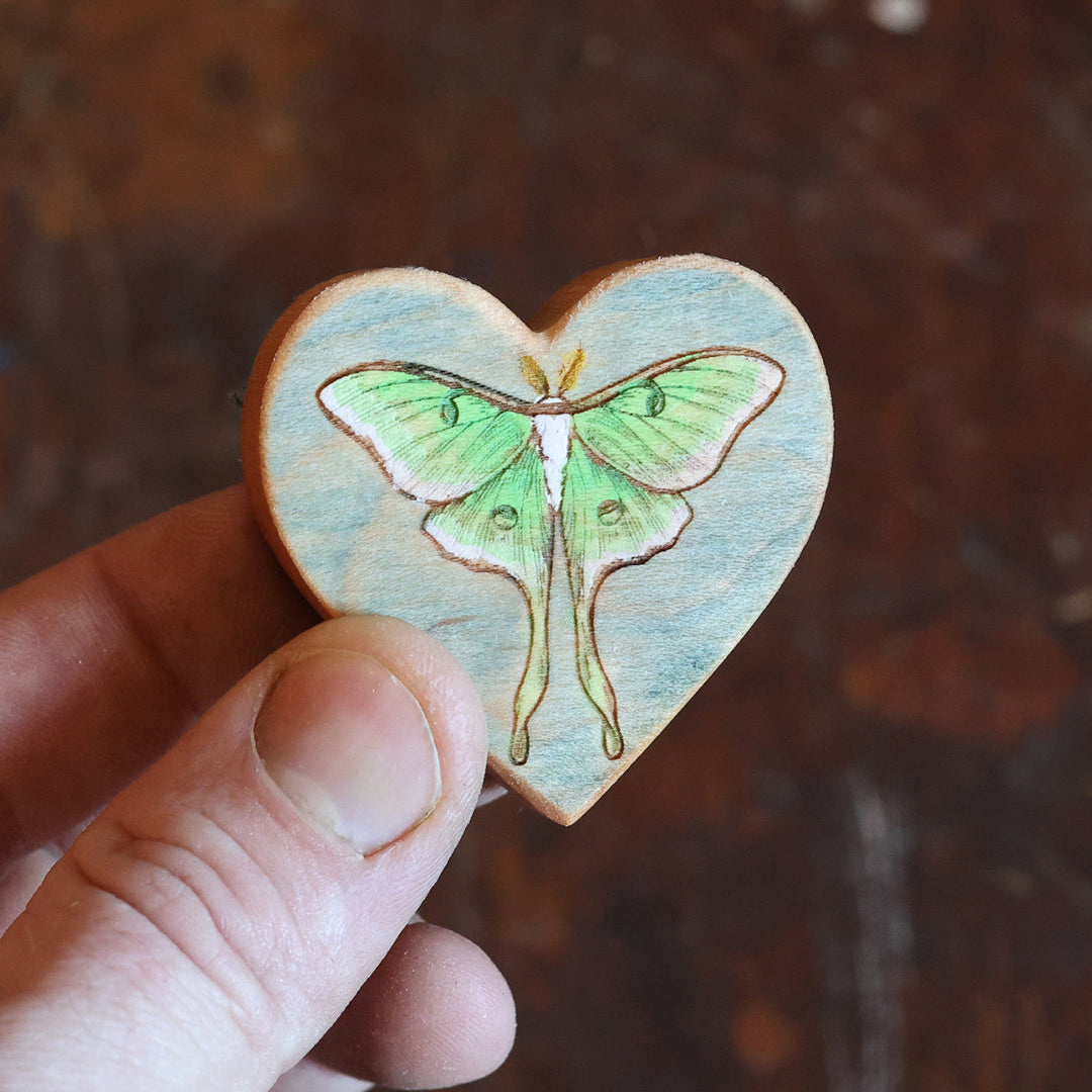 A hand holding a small heart-shaped wooden plaque featuring a painted green luna moth on a light blue background, set against a dark rustic wooden surface.